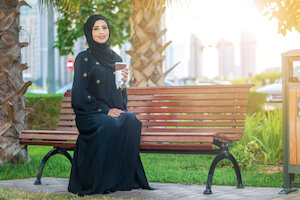 Smiling Arab woman dressed in an abaya sitting on a bench in sunshine - image by shutterstock.com